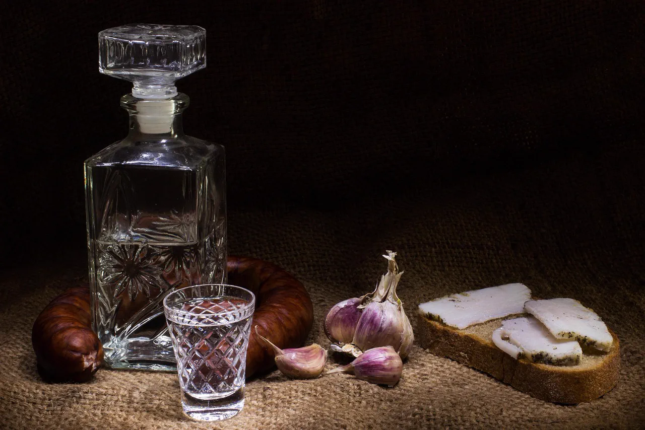 Carafă de cristal cu un păhărel, cârnat afumat, usturoi și pâine cu slănină, pe o pânză de sac
