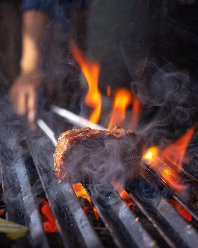 Bucată de carne întoarsă pe grătar, printre flăcări și fum